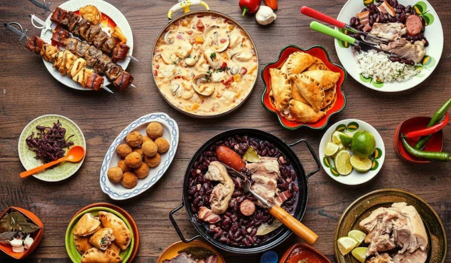 Uma vista aérea de uma mesa de madeira escura coberta com uma variedade de pratos tradicionais brasileiros e latino-americanos em tigelas e pratos coloridos.